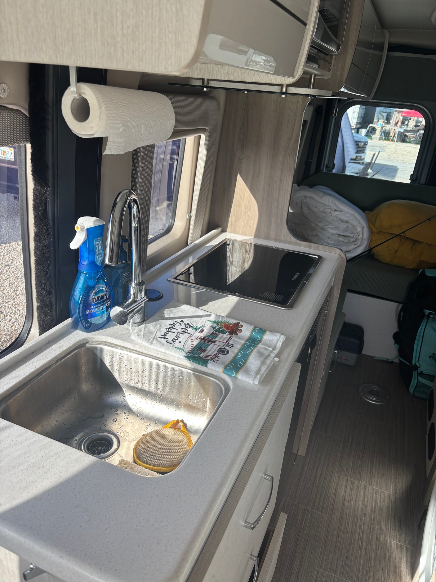 Compact RV rental kitchen features a sink, stove, Dawn Powerwash, and "Happy Camper" towel, ready for adventure.