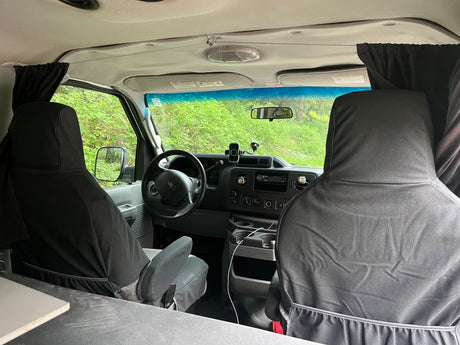 Interior view of an RV rental van: front seats, dashboard, steering wheel, and black privacy curtains, with green trees outside.