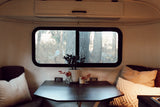 Inside a cozy RV rental: dinette with dark table, cushions, and a large window showcasing a natural view.
