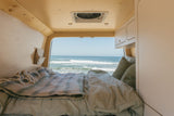 A cozy camper van bed faces an ocean view with waves, ideal for an RV rental getaway.