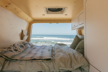 A cozy camper van bed faces an ocean view with waves, ideal for an RV rental getaway.