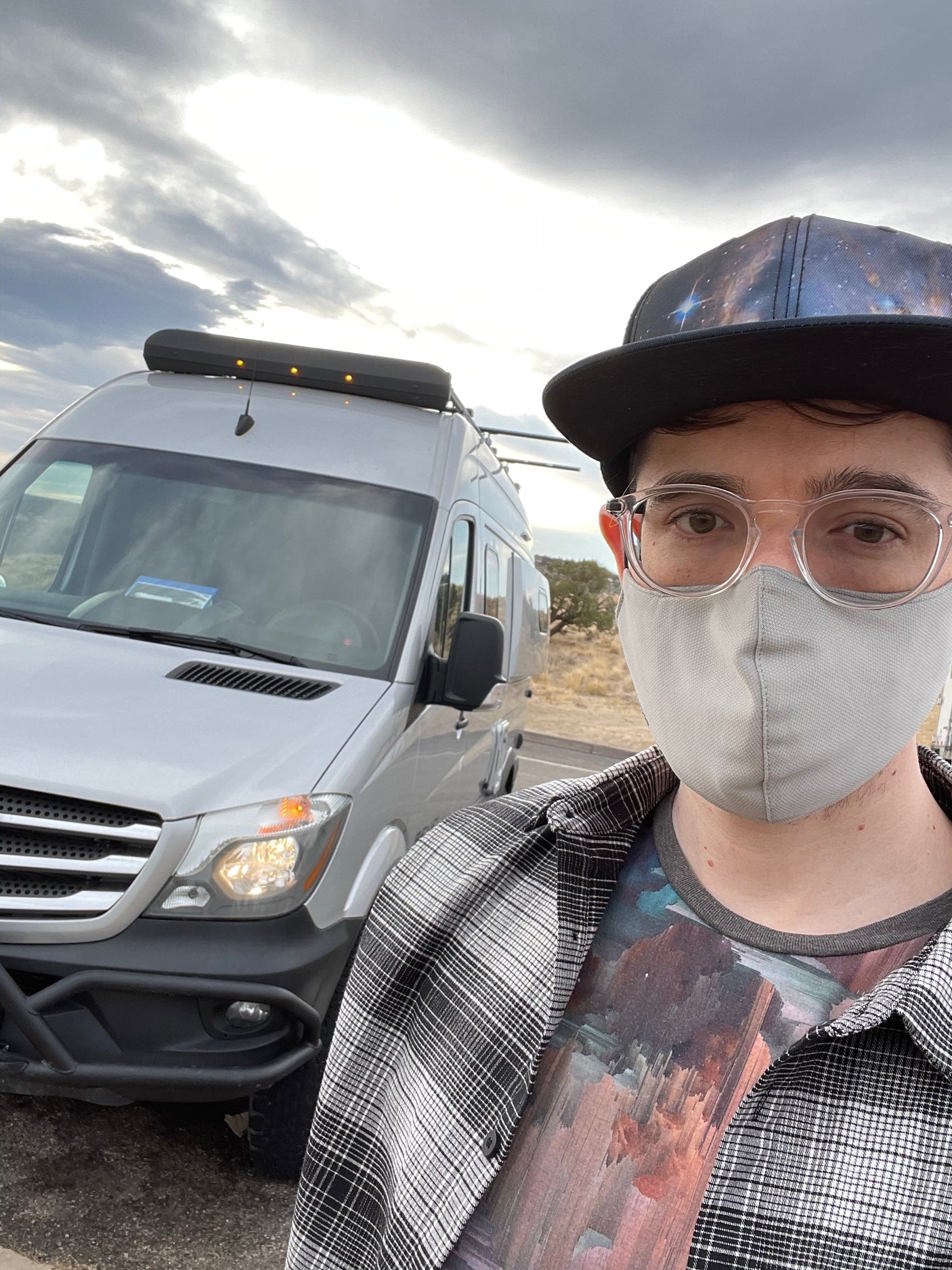 Selfie with an RV rental. A person in glasses, mask, and galaxy hat stands by a white campervan under a cloudy sky, ready for travel.