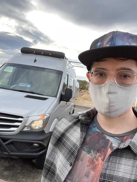 Selfie with an RV rental. A person in glasses, mask, and galaxy hat stands by a white campervan under a cloudy sky, ready for travel.