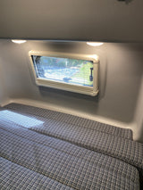 An RV rental's interior features a window with blinds, bright overhead lights, and patterned cushions set up as a comfortable bed.