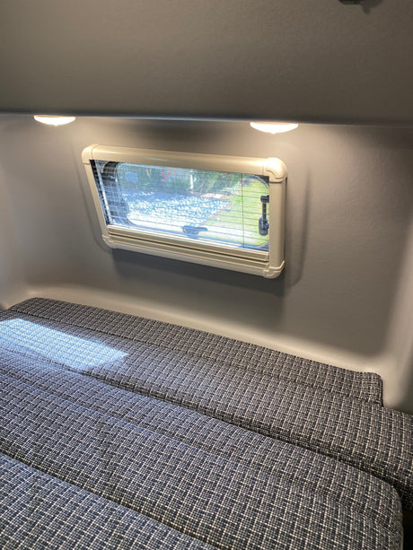 An RV rental's interior features a window with blinds, bright overhead lights, and patterned cushions set up as a comfortable bed.