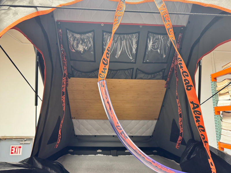 Interior of the Alu-Cab Gen 3-R rooftop tent looking toward the head end, showing the bamboo drop-down table panel, quilted insulated lining, clear-front storage pockets and orange Alu-Cab webbing straps — photo 5