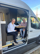 Inside a bright RV rental van, a smiling couple and their dog relax at a table, ready for travel.
