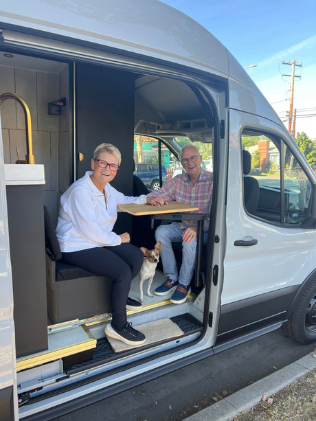 Inside a bright RV rental van, a smiling couple and their dog relax at a table, ready for travel.