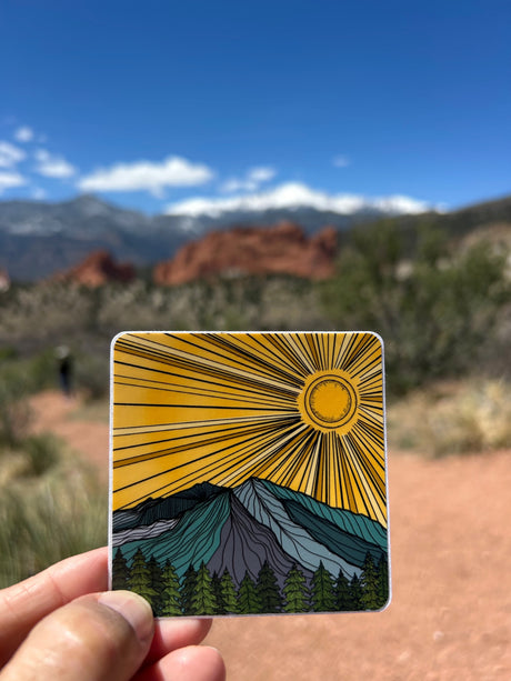 Hand holds a mountain & sun sticker, mirroring a blurred scenic mountain backdrop. Great for your next rv rental adventure!