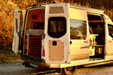 Spacious white camper van RV rental, rear and side doors open. Inside: bed, storage, table with wine. Ready for outdoor adventure.