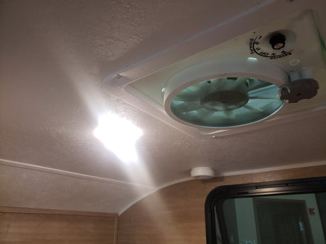 Overhead view in an RV rental: bright ceiling light, vent fan with controls, smoke detector, and window below.