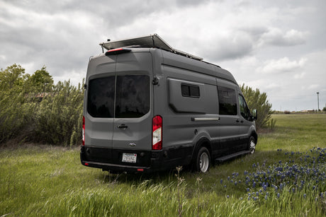 Gray Ford Transit camper van (RV rental) with solar panels parked in a grassy field with bluebonnets under a cloudy sky.