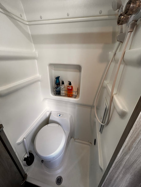 Inside an RV rental: a bright white, compact wet bath featuring a toilet, integrated shower, and toiletries shelf.