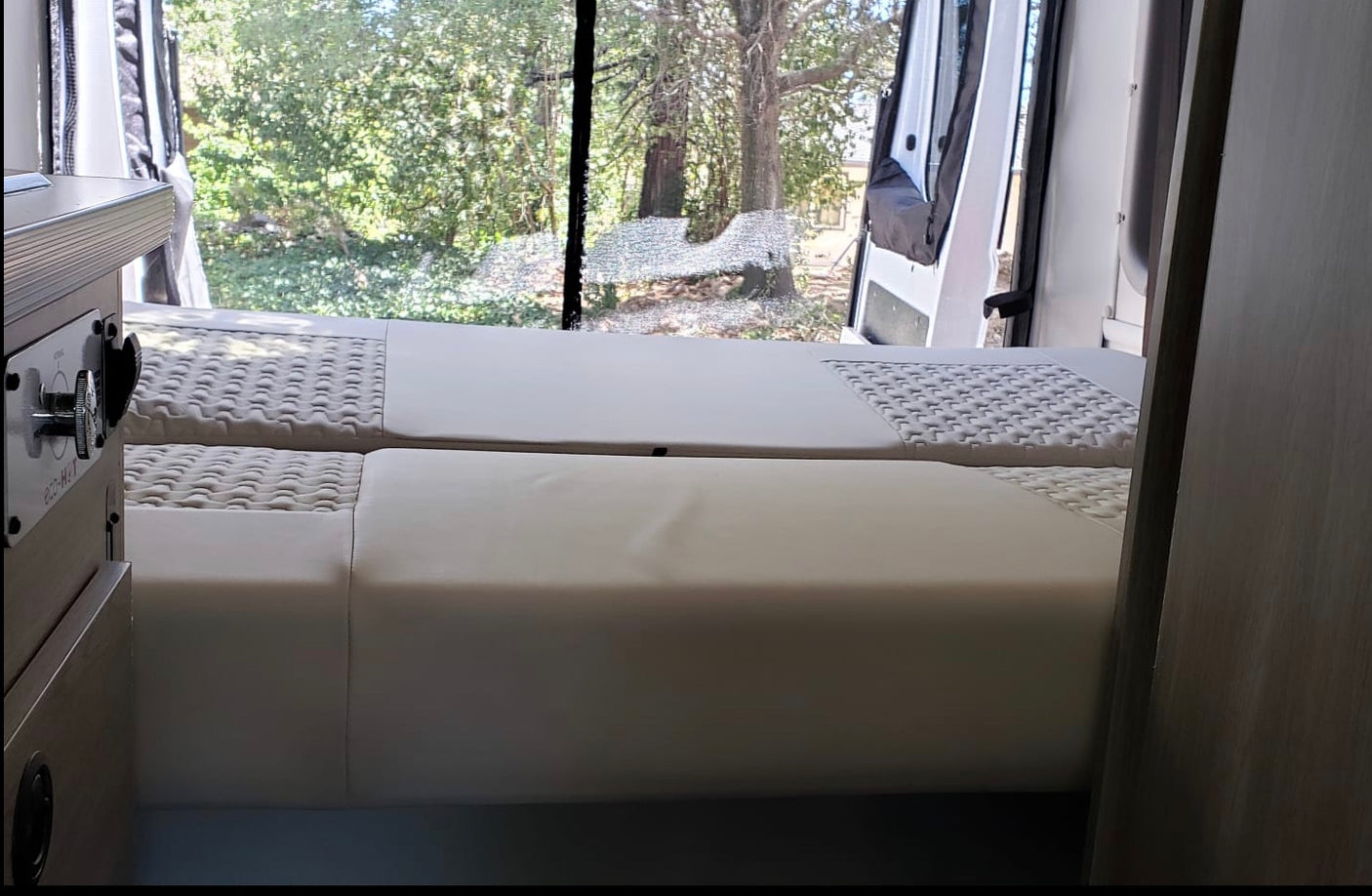 RV rental interior shows comfortable white beds. Rear doors are open, offering a scenic view of trees and nature outside.
