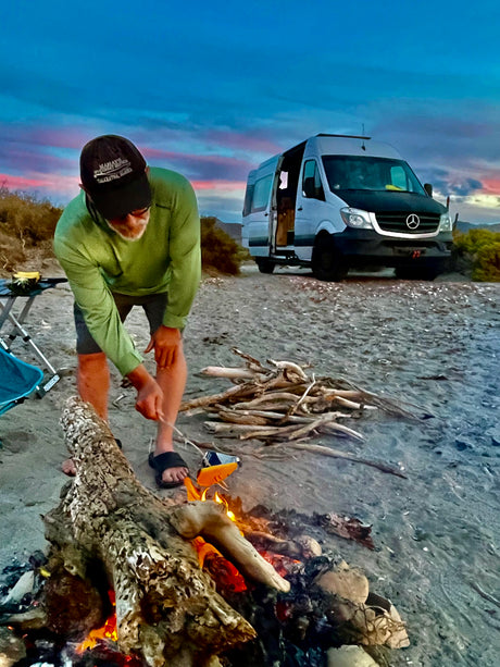 Man starting campfire by his RV rental camper van on a sandy beach at sunset, enjoying van life.