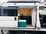 This white camper van rv rental opens to reveal a custom, wood-paneled interior with a kitchen, bed, and adventure-ready roof features.
