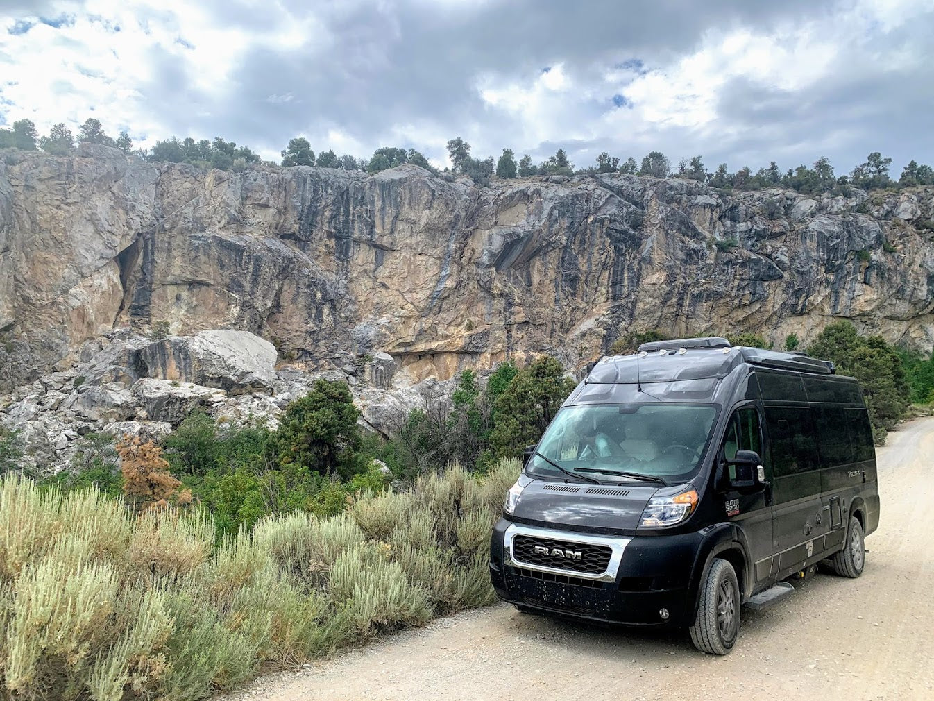Black Ram camper van, perfect as an rv rental, parked on a dirt road beside a towering rock cliff. Ideal for off-road adventures.