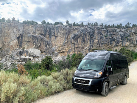 Black Ram camper van, perfect as an rv rental, parked on a dirt road beside a towering rock cliff. Ideal for off-road adventures.