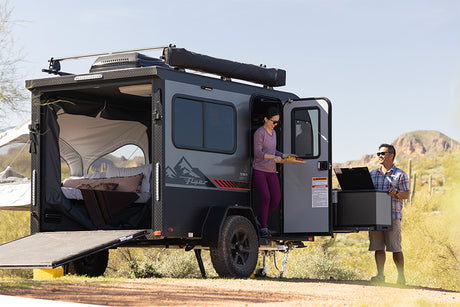Couple enjoys their compact RV rental, preparing food in a sunny desert landscape next to their unique travel trailer with open rear.