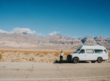 A traveler with arms raised stands beside a white camper van, an RV rental, on a desert road with mountains in the background.