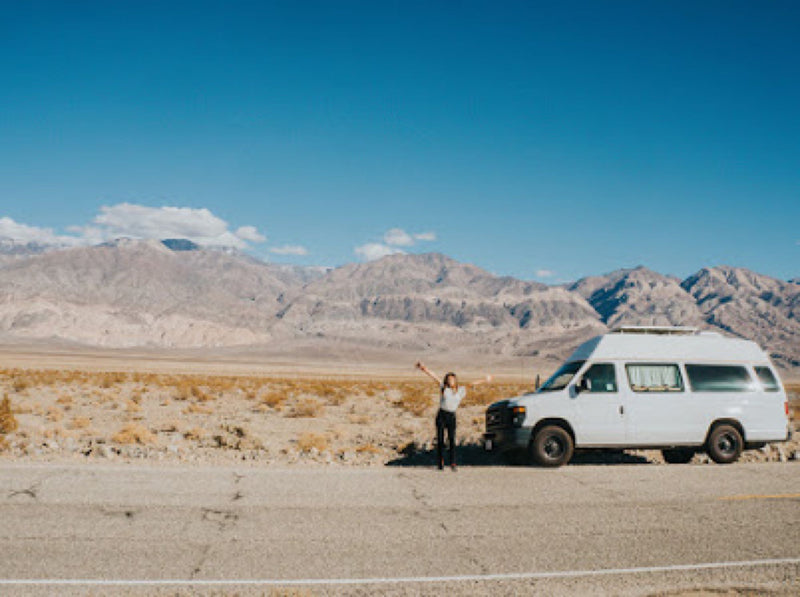 A traveler with arms raised stands beside a white camper van, an RV rental, on a desert road with mountains in the background. — photo 8