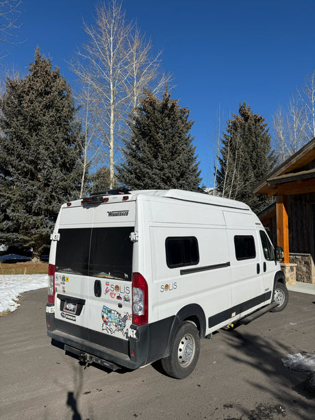 A white Winnebago Solis camper van, popular for rv rental, parked on an asphalt driveway with snow, surrounded by pine and bare trees.