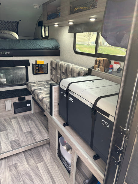 Compact rv rental interior with bed, patterned seating, storage, and a Dometic CFX cooler, offering a functional living space.