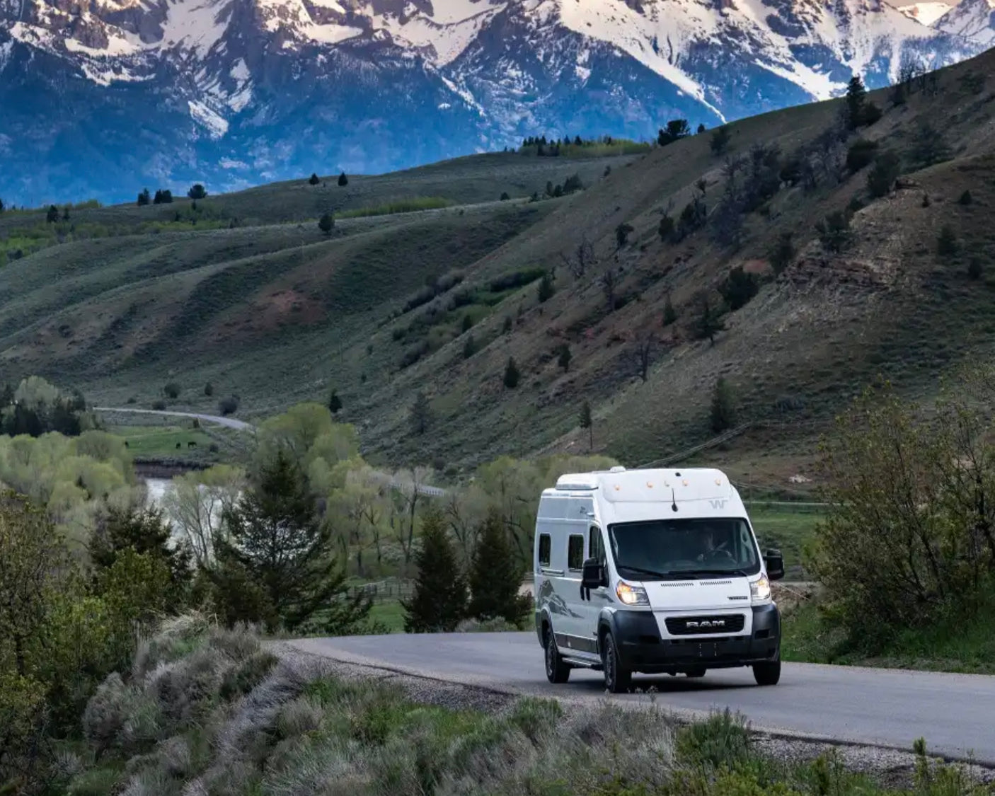 White rv rental van on a mountain road with a river and green hills. Ideal for scenic travel.