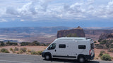 A white rv rental (Winnebago Solis) is parked by a road, offering a panoramic view of layered desert canyons and mesas under a cloudy sky.
