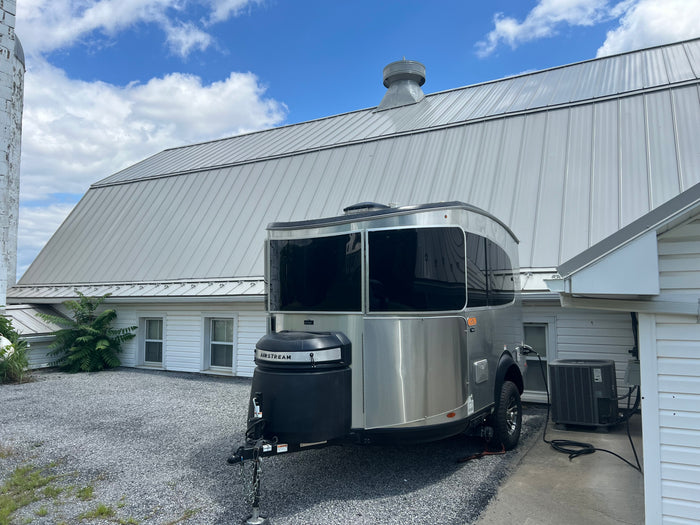An Airstream RV rental is parked on a gravel driveway next to a barn with a metal roof under a blue sky.