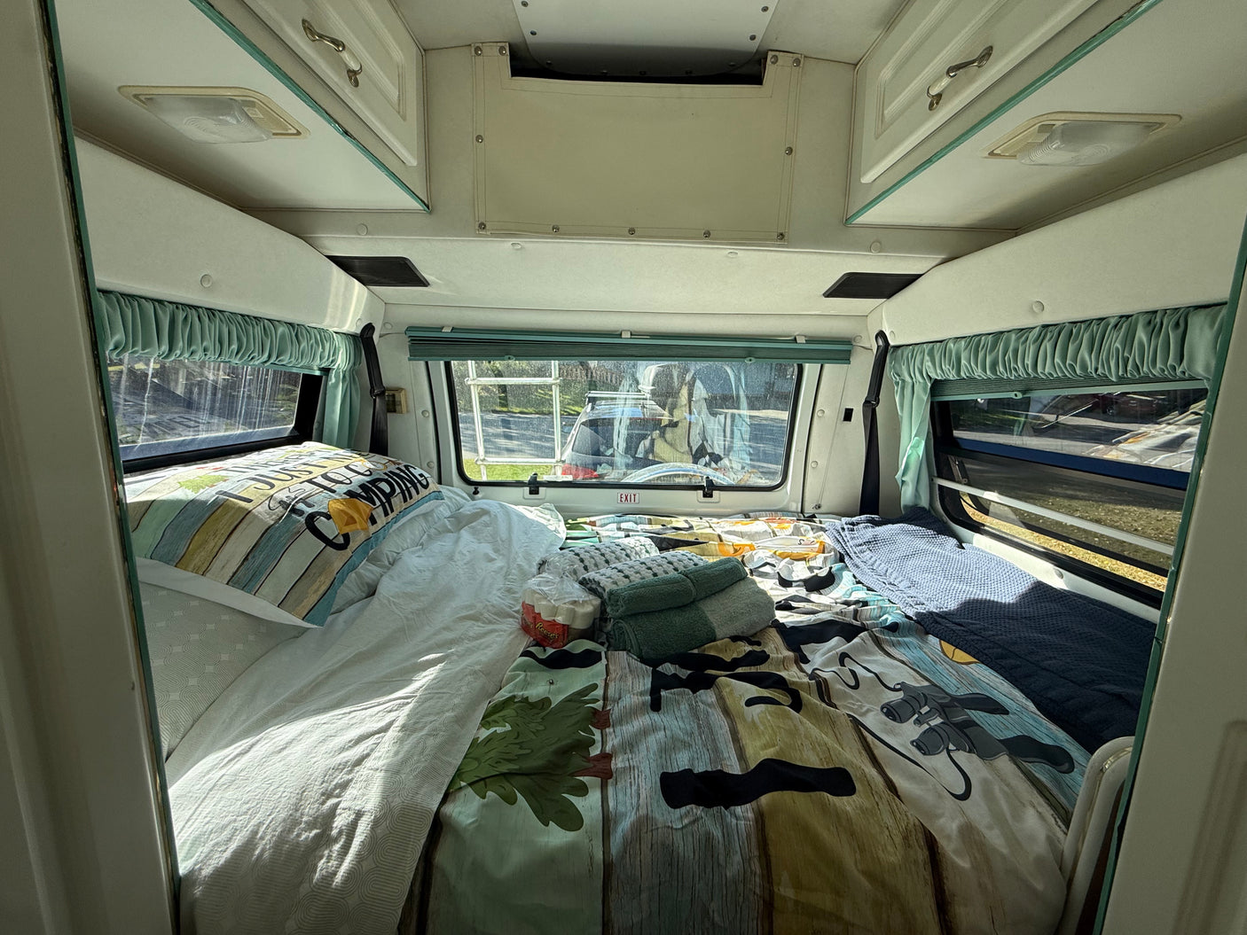 Full-Size Bed with Memory Foam Topper – Sleeps two adults comfortably after a day of exploring. Bed with camping comforter, pillows, windows with curtains, and overhead storage.
