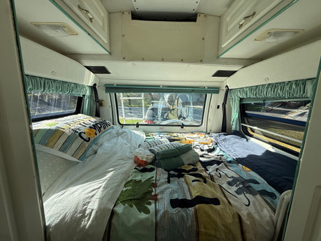 Full-Size Bed with Memory Foam Topper – Sleeps two adults comfortably after a day of exploring. Bed with camping comforter, pillows, windows with curtains, and overhead storage.