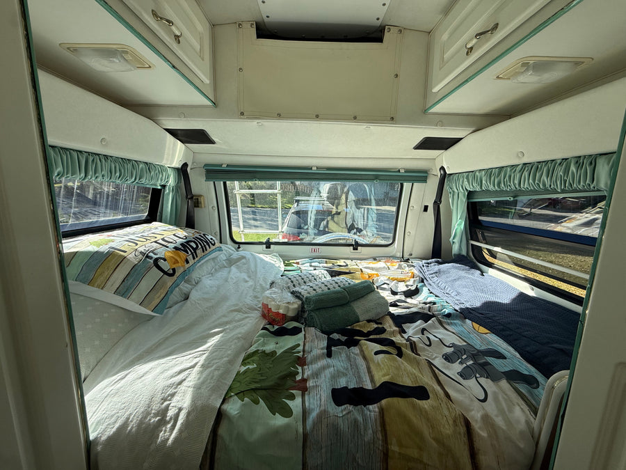 Full-Size Bed with Memory Foam Topper – Sleeps two adults comfortably after a day of exploring. Bed with camping comforter, pillows, windows with curtains, and overhead storage.