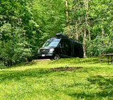 Black Mercedes Sprinter RV rental van at a lush green campsite with a picnic table, surrounded by trees.