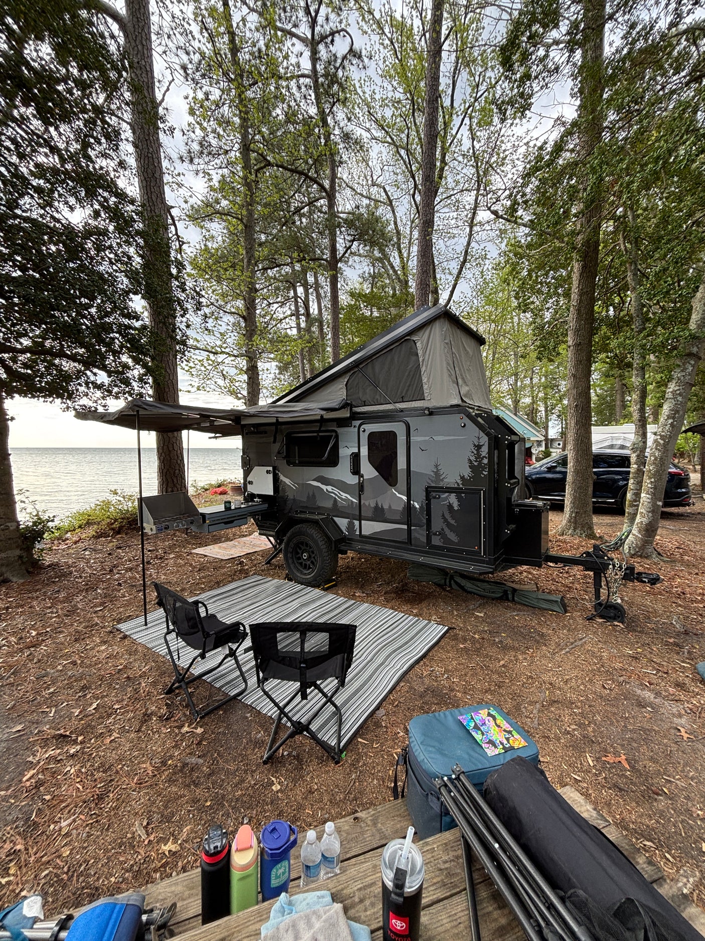 Waterside RV rental setup: a pop-up camper with awning, outdoor kitchen, chairs, and gear, nestled by the lake.