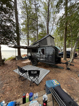 Waterside RV rental setup: a pop-up camper with awning, outdoor kitchen, chairs, and gear, nestled by the lake.