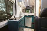 Inside a modern rv rental: compact camper van kitchen with sink, induction cooktop, fridge, and cozy bed in the rear.