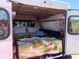 Cozy RV rental interior: bed with plaid and "Home Sweet Home" blanket, laptop, and upper storage shelves. Ideal for travel.