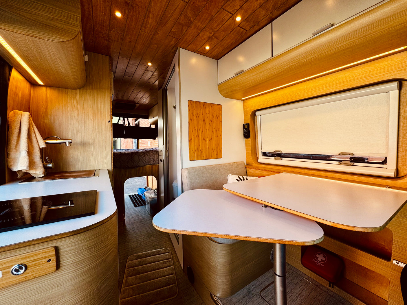 Cozy RV rental interior with a compact kitchen, dining table, and warm wood accents. A bed area is visible in the back.
