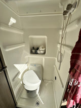 Inside a compact RV rental wet bath with toilet, shower, and shelving. A red towel hangs, and surfaces are wet.