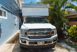 White Ford truck with a camper shell, likely an RV rental, parked on a paved driveway in Hawaii, between a blue house and lush plants.