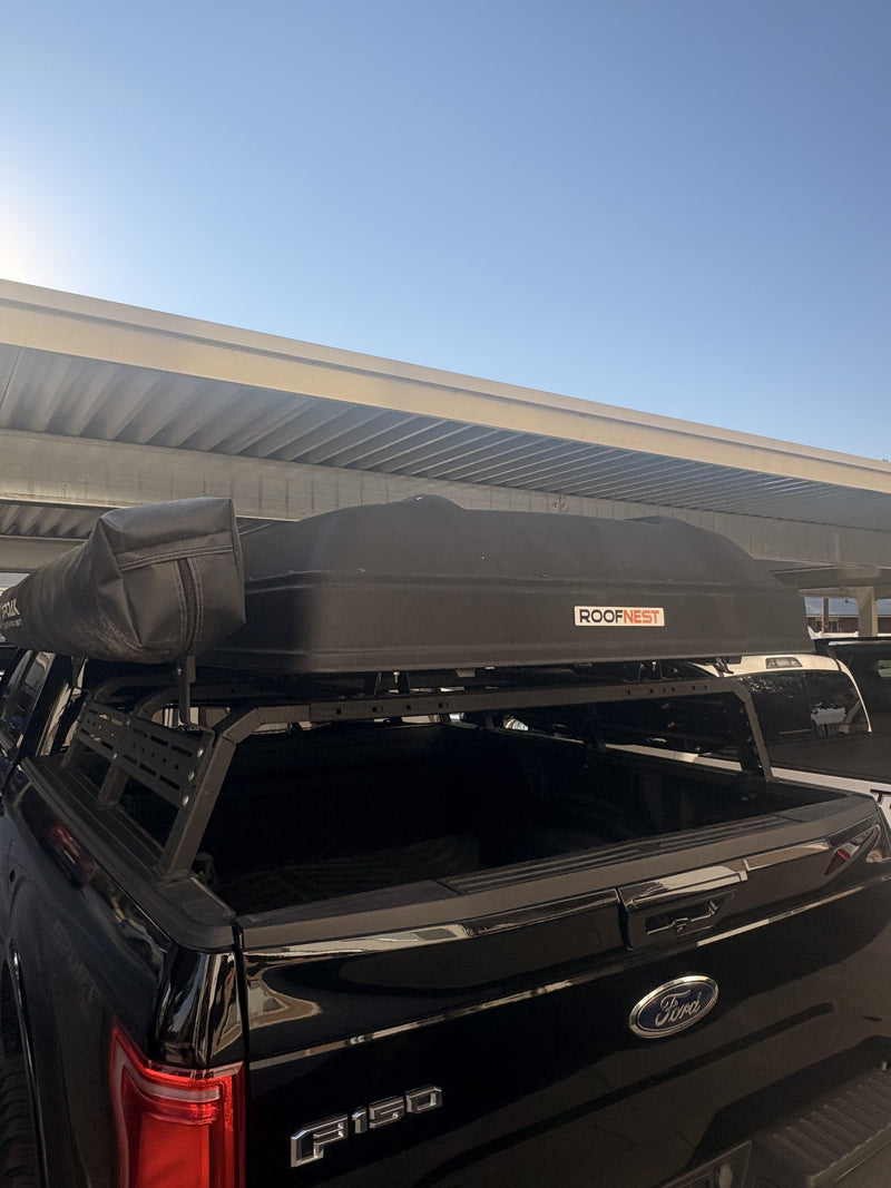 Roofnest Condor XL hardshell rooftop tent closed on an RCI bed rack over a black Ford F-150, ROOFNEST badge visible on the shell and the rolled Topoak awning bag mounted alongside — photo 1