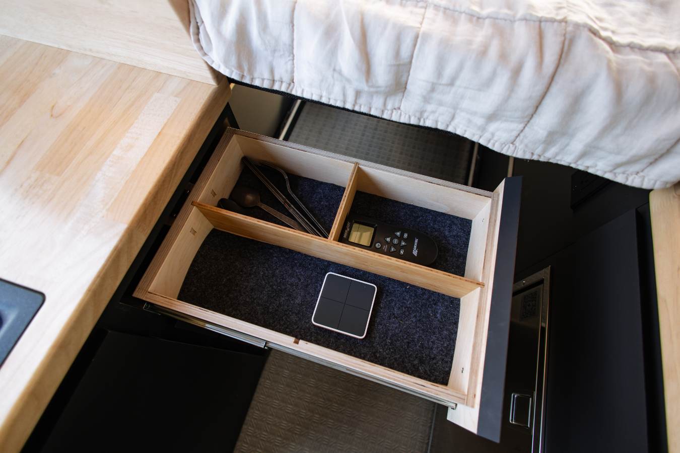 RV rental storage: an open drawer with utensils, a remote, and a control panel, next to a bed and wooden counter.