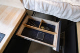 RV rental storage: an open drawer with utensils, a remote, and a control panel, next to a bed and wooden counter.