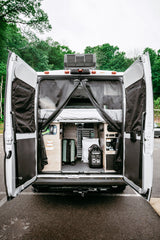 Inside an RV rental camper van: rear doors open, revealing organized storage, a bed, a suitcase, and a Powerhouse generator, ready for adventure.