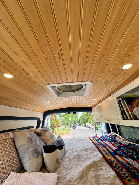 Inside a cozy RV rental: wooden ceiling, fan, and bed looking out to a tree-lined street with parked cars.