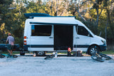 Adventure-ready RV rental setup: white camper van with awning, bikes, and a person unpacking in a wooded area.
