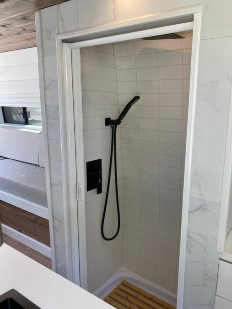 Modern RV rental bathroom with sleek white tiled shower, black fixtures, and a wooden floor mat for comfortable travel.