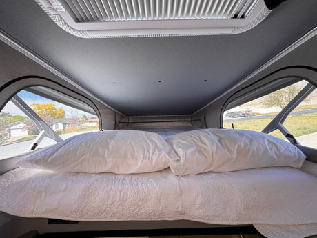Cozy rv rental bed with white pillows and a quilted comforter, offering views of houses and trees through pop-top windows.