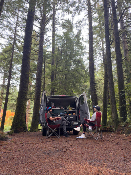 Two people relax by their **rv rental** van, ready for camping in a serene forest with tall trees. Perfect for outdoors.